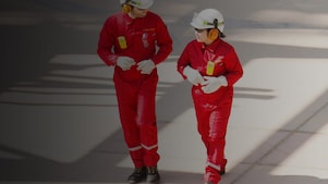 Un trabajador y una trabajadora en overol rojo y casco blanco, con protectores auditivos, caminando