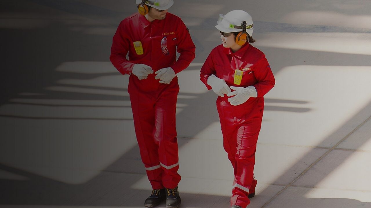 Un trabajador y una trabajadora en overol rojo y casco blanco, con protectores auditivos, caminando
