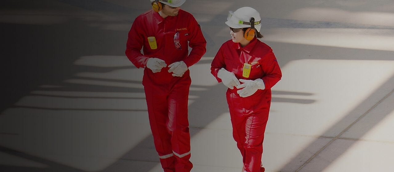Un trabajador y una trabajadora en overol rojo y casco blanco, con protectores auditivos, caminando