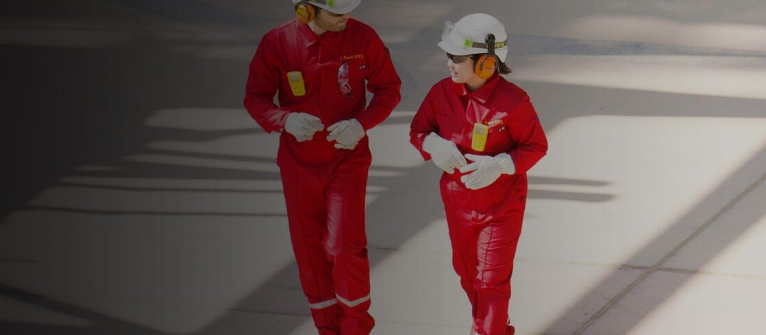 Un trabajador y una trabajadora en overol rojo y casco blanco, con protectores auditivos, caminando