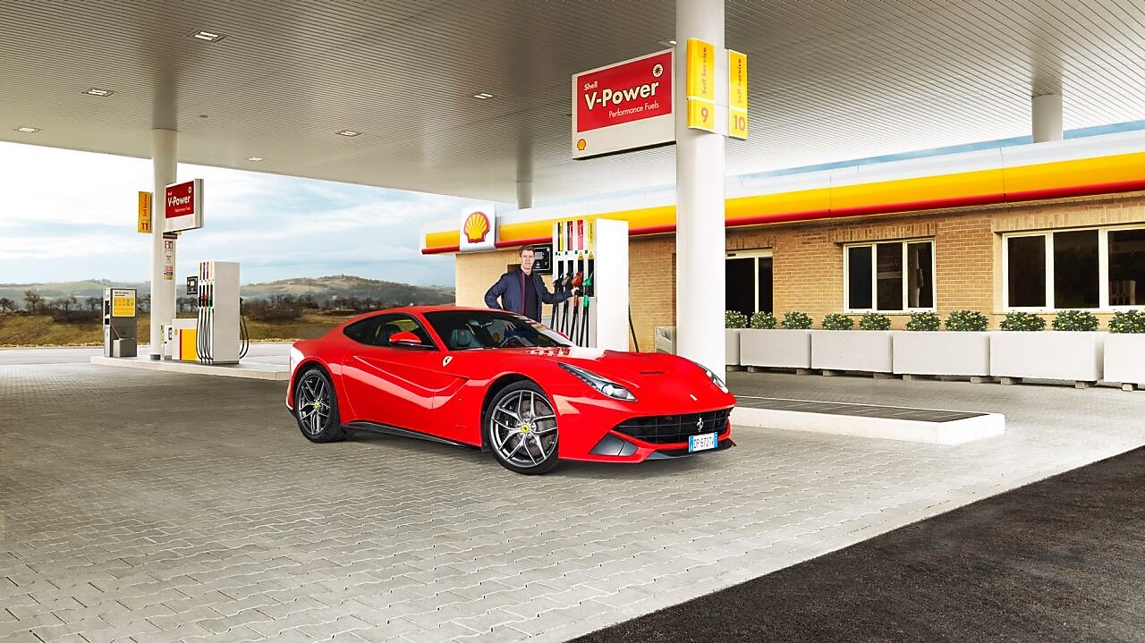 Ferrari rojo en una estación de servicio Shell con un hombre apoyado sobre un surtidor
