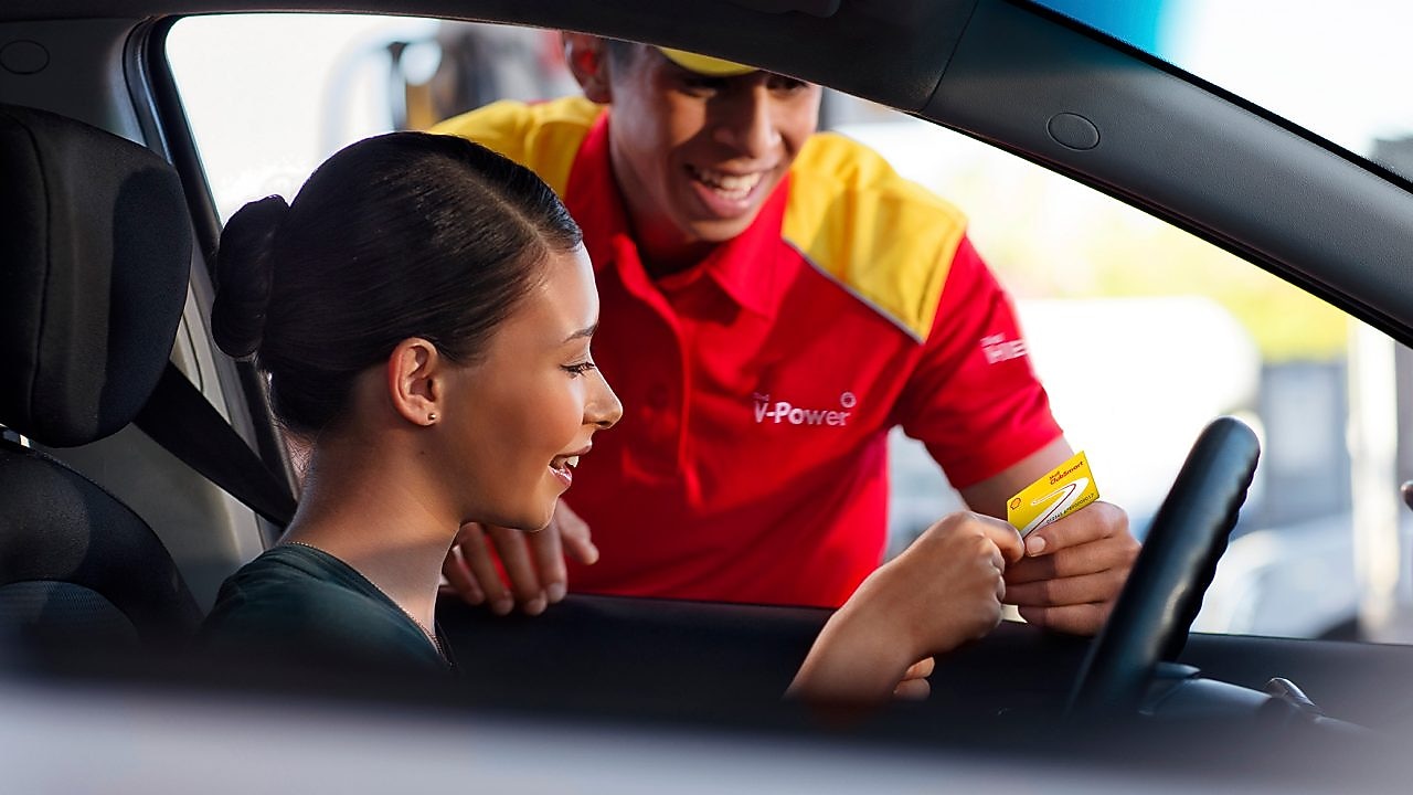 Mujer al volante, en un coche parado, siendo atendida por un empleado de una gasolinera Shell. Tiene el pelo negro y sonríe, lleva uniforme y una gorra roja. Ella le enseña una tarjeta Shell.